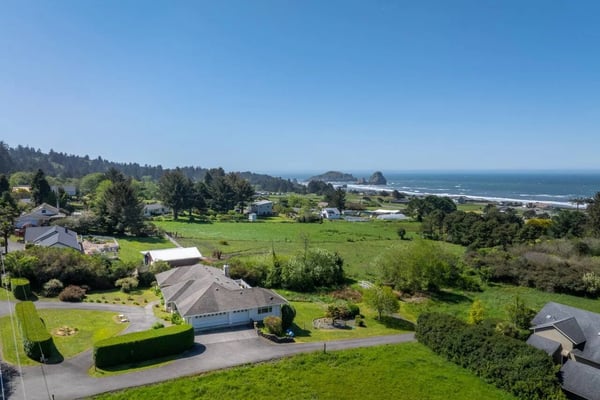 Aerial view of rural coastal property with house, green pastures, forested hills, and ocean in distance