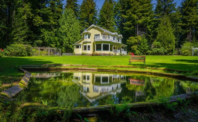 Yellow farmhouse with wraparound porch surrounded by tall evergreen trees, green lawn, and small pond with reflection in foreground.